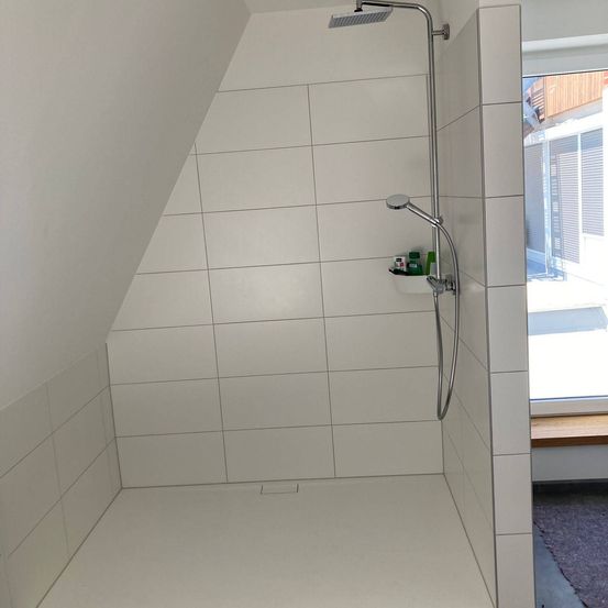 Ein Duschbereich im Dachgeschoss mit weißen Fliesen, einer Duschbrause und einem Fenster, das ein Haus und einen Garten zeigt.