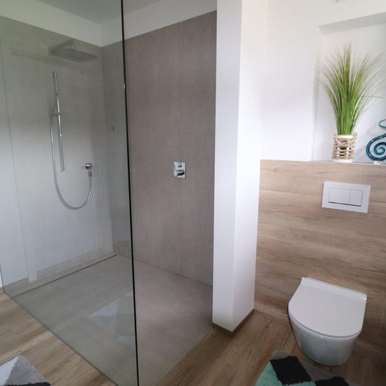 Ein modernes Badezimmer mit einer Glasdusche, einem weißen WC und einer Holzwand. An der Wand steht eine Pflanze in einem Korb.