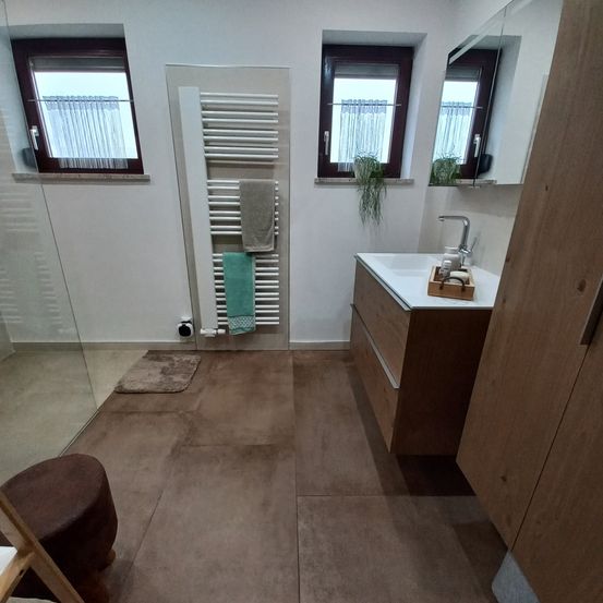 Ein Badezimmer mit einer Glasdusche, braun gefliestem Boden und einem Waschbecken mit Wasserhahn. Es gibt zwei Fenster, eines mit einer Pflanze und das andere mit einem weißen Vorhang. Ein Handtuchhalter mit Handtüchern hängt an der Wand.