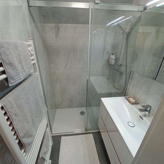 Ein modernes Badezimmer mit einer gläsernen Duschkabine, einem weißen Waschbecken und einem Handtuchhalter mit Handtüchern. Ein Spiegel reflektiert den Raum, und eine Seifenschale ist auf dem Waschbecken.