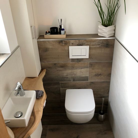 Ein kleines Badezimmer mit Holzboden und weißen Wänden. Ein weißes WC steht neben einem Waschbecken mit Wasserhahn. Ein Tablett mit Flaschen und einer Pflanze befindet sich auf dem Holzregal über dem WC.