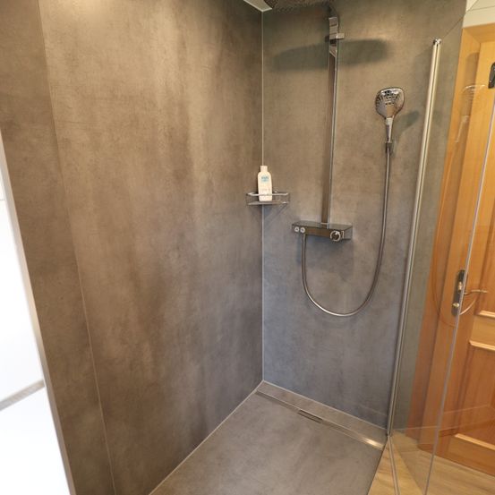 Ein moderner Duschbereich mit grau strukturierten Wänden, einer Glastür und einer an einer silbernen Halterung befestigten Duschkopf. Ein kleiner Regalboden hält eine Flasche.