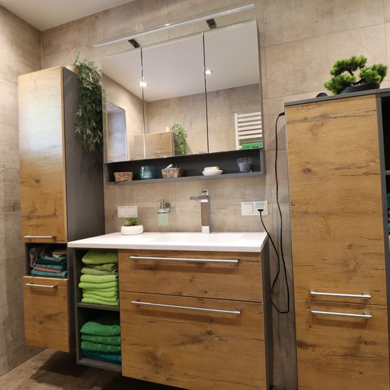 Ein modernes Badezimmer mit Holzschränken, einem weißen Waschbecken, einem Spiegel, einer Topfpflanze und einem Regal mit verschiedenen Gegenständen.