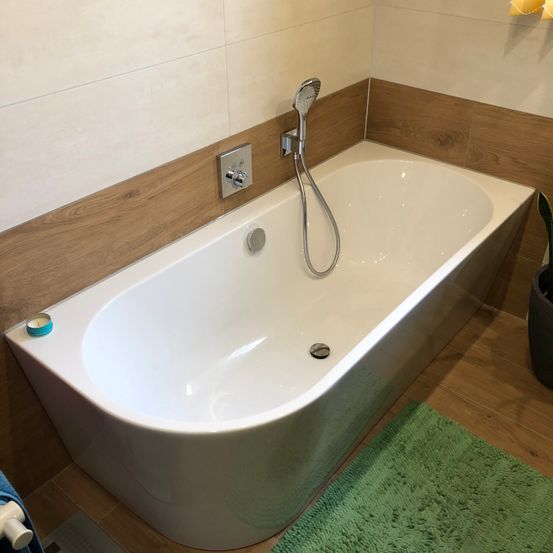 Eine moderne weiße Badewanne mit einem Duschkopf befindet sich in einem Badezimmer. Sie ist von Holzwänden umgeben und hat einen gefliesten Boden. Ein grünes Handtuch liegt auf dem Boden.