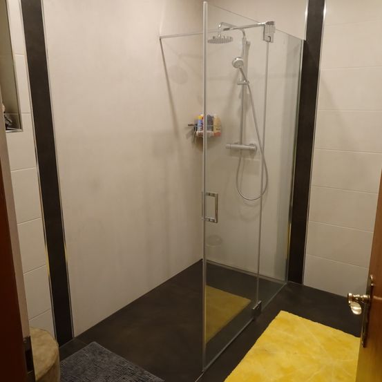 Eine Glasdusche mit silbernem Griff, Duschkopf und Regal mit Gegenständen. Der Boden ist mit schwarzen Fliesen belegt, und auf dem Boden liegt ein gelber Teppich. Die Wände sind mit weißen Fliesen und schwarzem Rand versehen.