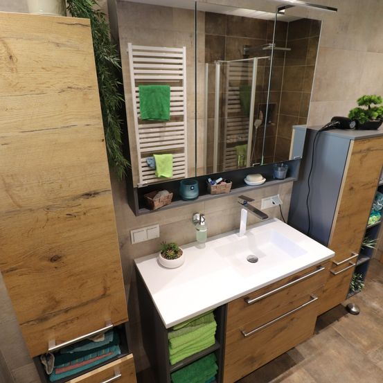 Ein Badezimmer mit Holzschränken, einem weißen Waschbecken, einem Spiegel und einer Glasdusche. Die Regale halten Handtücher, Seife und Pflanzen.