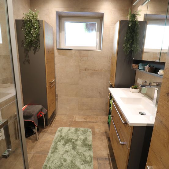 Ein modernes Badezimmer mit gefliesten Wänden, Holzschränken, einem weißen Waschbecken, einem Spiegel und einem Fenster. Ein grüner Teppich liegt auf dem Boden.