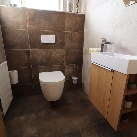 Ein Badezimmer mit braunen Fliesen, einer weißen Toilette, einem Waschbecken mit Wasserhahn und einem Holzschrank mit Regalen sowie einem weißen Heizkörper.