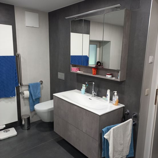 Ein modernes Badezimmer mit einem großen Spiegel über einem Waschbecken, einer Toilette und einem Handtuchhalter mit einem blauen Handtuch. Die Wände sind grau und in der Nähe befinden sich ein Mülleimer und eine Waage.