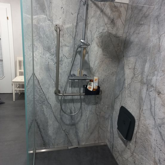Eine moderne Duschkabine mit einer klaren Glastür, grauen Marmorfliesen, einer silbernen Duschbrause und einem schwarzen Seifenhalter mit Toilettenartikeln.