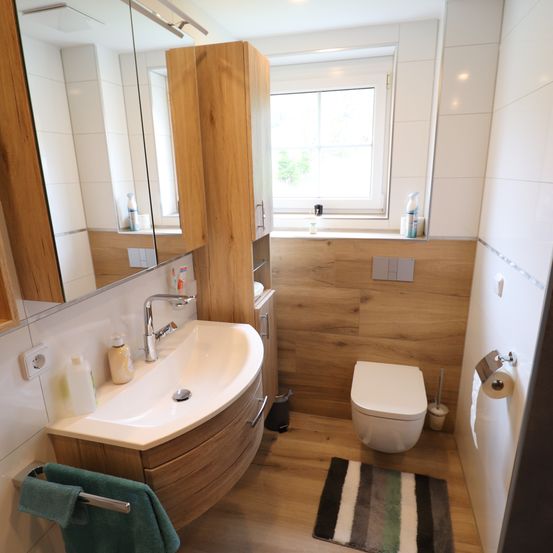 Ein Badezimmer mit Holzboden und -wänden, mit einem Waschbecken mit Wasserhahn, einer Toilette und einem Fenster. Ein Spiegel hängt an der Wand und ein Handtuch hängt an einem Halter.