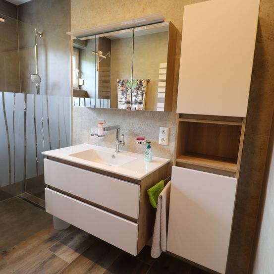 Ein Badezimmer mit einem weißen Waschbecken, einem Spiegel, Holzschränken und einer Glasdusche. Ein Handtuch hängt an der Wand.