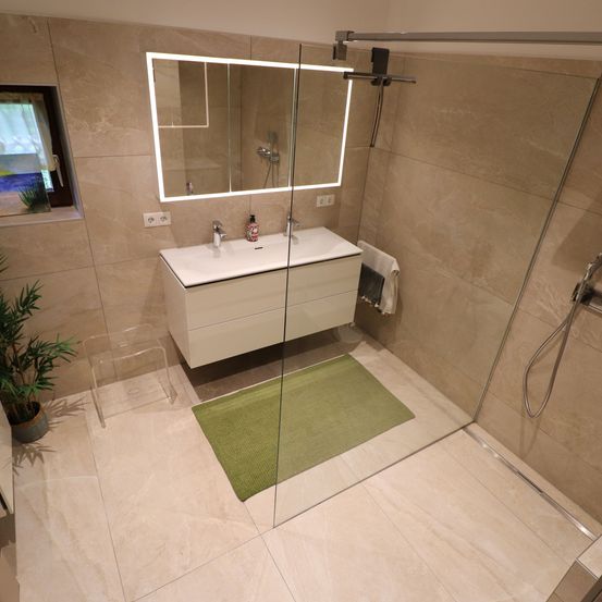 Ein Badezimmer mit einer Glasdusche, einem weißen Waschbecken, einem Spiegel und einem grünen Teppich. Eine Pflanze und ein Fenster sind ebenfalls vorhanden.