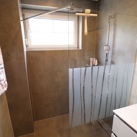 Ein Badezimmer mit einer Glasdusche, einem Fenster und einer Duschbrause mit Schlauch. Die Dusche hat einen Sichtschutz mit vertikalen Streifen.