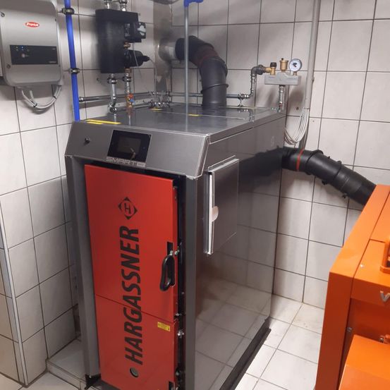 Eine große, rote und silberne Maschine mit dem Wort 'HARGASSNER' darauf, an einer weißen gefliesten Wand montiert. Sie hat ein digitales Display, angeschlossene Rohre und einen großen Abgasrohr.
