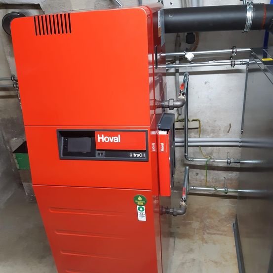 Ein roter Hova-Kessel mit einem Ultra Oil-Aufkleber steht in einem Versorgungsraum, umgeben von Metallrohren und Schläuchen.