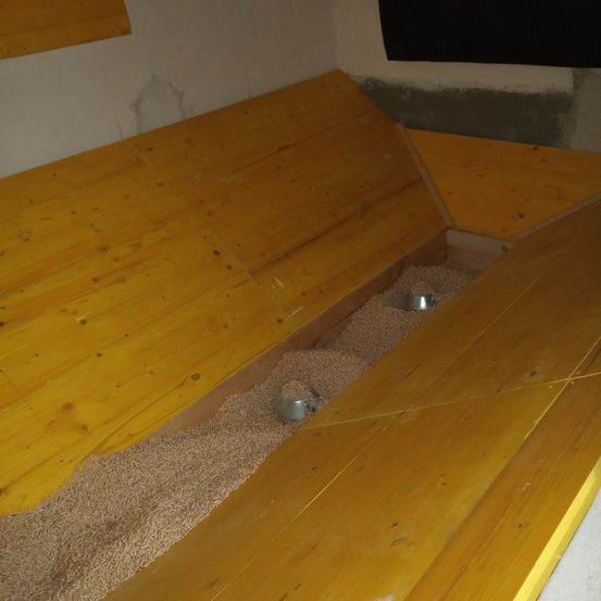 Ein Raum mit gelben Holzwänden und einem Holzboden, bedeckt mit einer Schicht beigefarbener Körner. Zwei Metallschalen sind in den Körnern platziert.