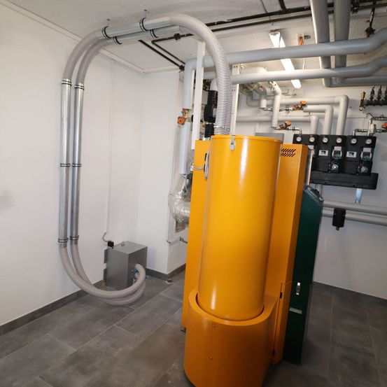 Ein Industrieraum mit weißer Wand und grau gefliestem Boden zeigt große gelbe und grüne Maschinen mit zahlreichen Rohren und Schläuchen.