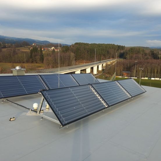 Ein Dach mit installierten Solarpanelen unter blauem Himmel. In der Ferne ist eine Brücke zu sehen, und die Gegend ist von Bäumen und Bergen umgeben.