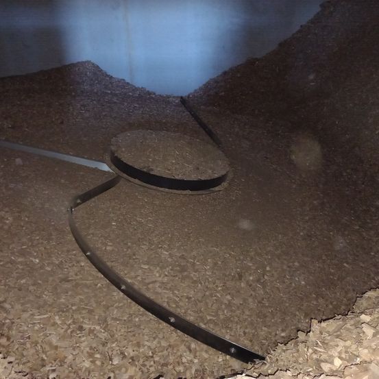 Ein dunkler Raum mit einem Haufen Holzspäne, einer runden Metallplatte und einem weißen Rohr liegt auf dem Boden. Der Raum hat eine blaue Wand.