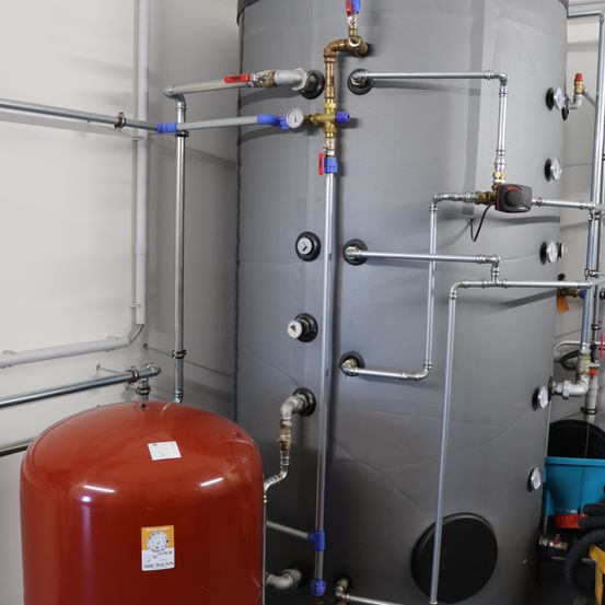 Ein Industrieraum mit einem großen grauen zylindrischen Speichertank, mehreren Rohren und Messgeräten sowie einem roten Tank mit Aufkleber.