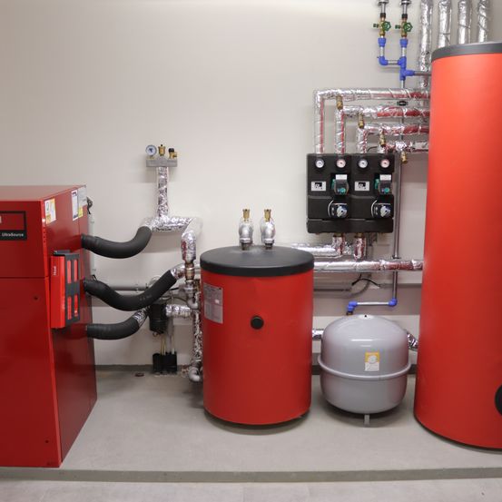 Ein Raum mit Industriegeräten, darunter ein roter Heizkörper, ein grauer Tank und ein roter Vorratstank, an einer weißen Wand mit Metallrohren montiert.