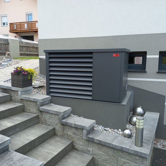 Eine graue Lüftungseinheit ist an der Seite eines Hauses installiert. Sie befindet sich auf den Stufen, mit einem Blumenkorb auf der linken Seite. Hinter dem Haus befindet sich ein Balkon und es gibt einen Zaun.