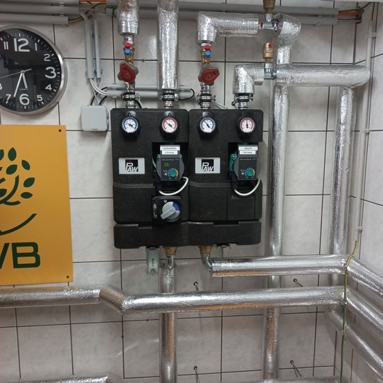 Industrielles Bedienfeld mit Manometern und Zählern an einer gefliesten Wand montiert. In der Nähe befindet sich ein gelbes Schild mit grünen Blättern und den Buchstaben VB. Eine Uhr zeigt die Zeit 11:55 an.