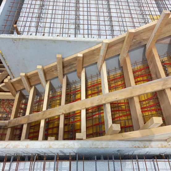 Eine Baustelle mit hölzernen Stützen und Stahlbewehrungsstäben. Die Holzbalken sind in einem V-förmigen Muster angeordnet. Die Stahlstäbe sind mit roten Drähten verbunden.