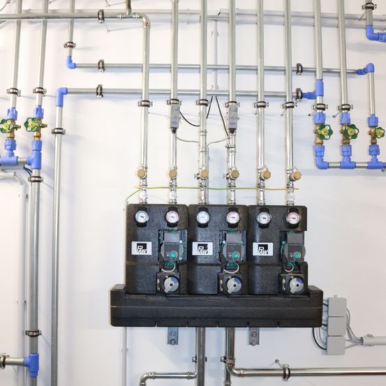 Eine Reihe von industriellen Steuereinheiten und Ventilen ist an einer weißen Wand montiert. Jede Einheit hat Manometer und Schalter, verbunden durch blaue und silberne Rohre.