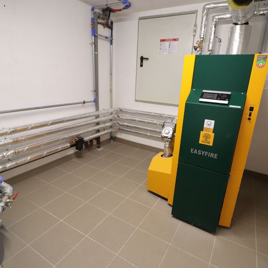 Eine gelbe und grüne Maschine mit der Aufschrift 'EASYFIRE' steht in einem Raum mit gefliesten Böden. Links sind Rohre an der Wand befestigt. Rechts ist eine weiße Tür teilweise geöffnet.