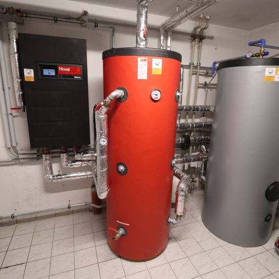 Ein Innenbereich mit zwei Wasserspeichern, einem roten und einem grauen, verbunden durch Rohre. Ein schwarzes Bedienfeld mit digitalem Display ist an der Wand montiert.