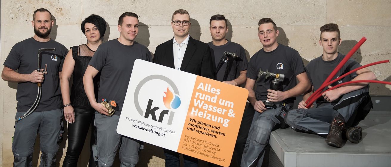 Fünf Männer posieren für ein Foto mit einem Schild. Vier halten Werkzeuge und Kameras, einer hält ein Schild mit 'KRI'. Das Schild hat ein Flammenlogo und steht 'Alles rund um Wasser & Heizung'. Sie tragen graue Hemden.