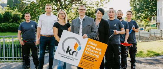Fünf Personen stehen nebeneinander und halten ein Schild mit einem Firmenlogo. Sie lächeln für ein Foto. Das Schild trägt den Text 'KRI Installationstechnik GmbH'.