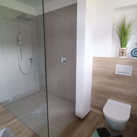 Ein modernes Badezimmer mit einer Glasdusche, Holzboden und einer weißen Toilette. Ein Topf mit einer Pflanze steht auf einem Regal über der Toilette.