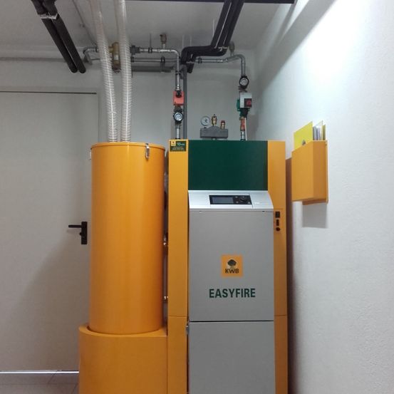 Ein gelber und grüner EASYFIRE Holzpelletkessel ist an einer weißen Wand montiert. Darauf ist ein Metallrohr an der Decke befestigt.