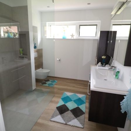 Ein Badezimmer mit Holzboden, einer Glasdusche, einer weißen Toilette und einem Waschbecken mit Armatur. Es gibt zwei Teppiche, einen Spiegel und ein Fenster über der Toilette.