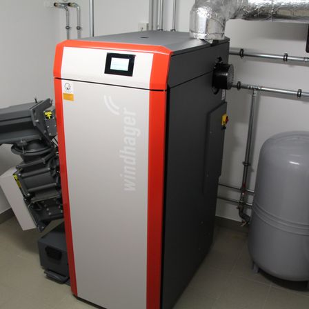Ein Windhager Heizsystem steht in einem Raum mit weißen Wänden, begleitet von Rohren und einem grauen Tank. Das System ist orange und grau mit einem digitalen Display auf der Vorderseite.
