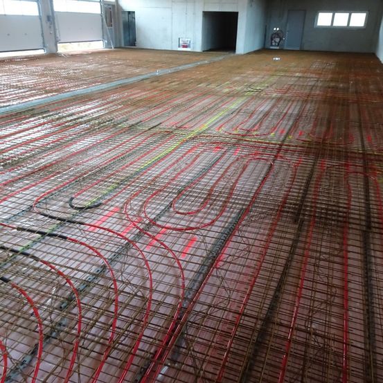 Ein leerer Raum mit unfertigen Böden zeigt ein Netzwerk von roten, gelben und grünen Rohren auf einem Drahtgitter. Große offene Türen und Fenster sind an den Wänden sichtbar.