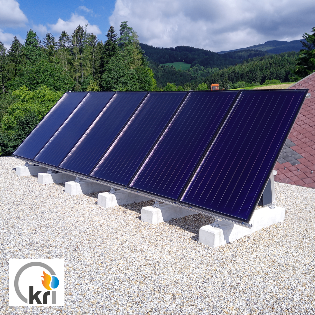 Six solar panels are installed on a roof. They face the sun, surrounded by trees and mountains. A logo with the text KRI is in the bottom left corner.
