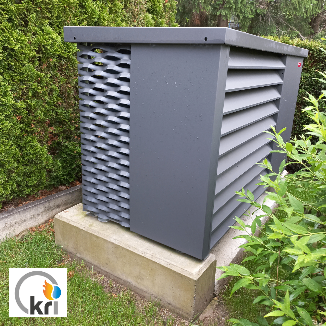 Eine graue Außenluftbehandlungseinheit mit Gitterdesign steht auf einem Betonblock in einem Garten mit grünen Büschen.
