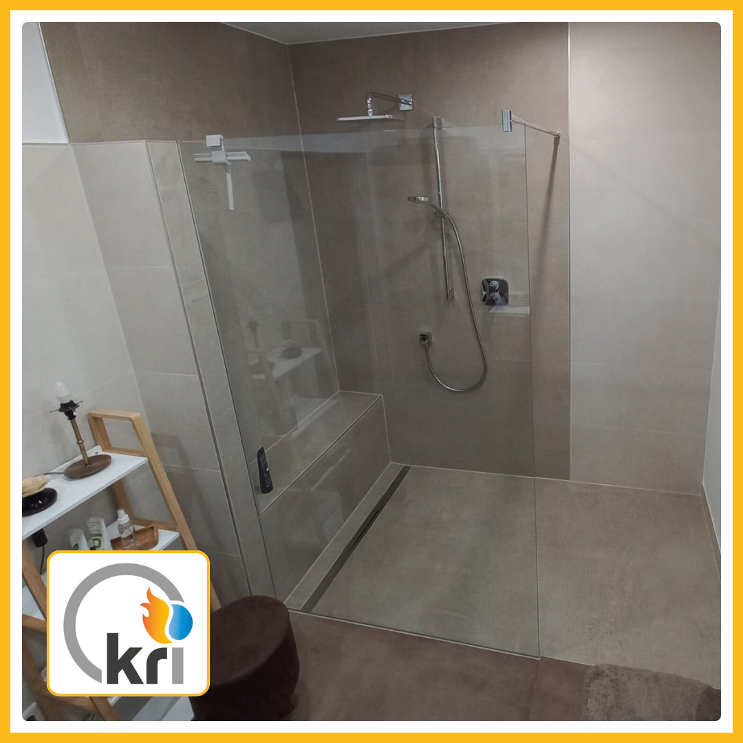 A modern bathroom with a glass shower enclosure, tiled floor, and a small shelf with a lamp and items. The shower has a silver handle and a showerhead.