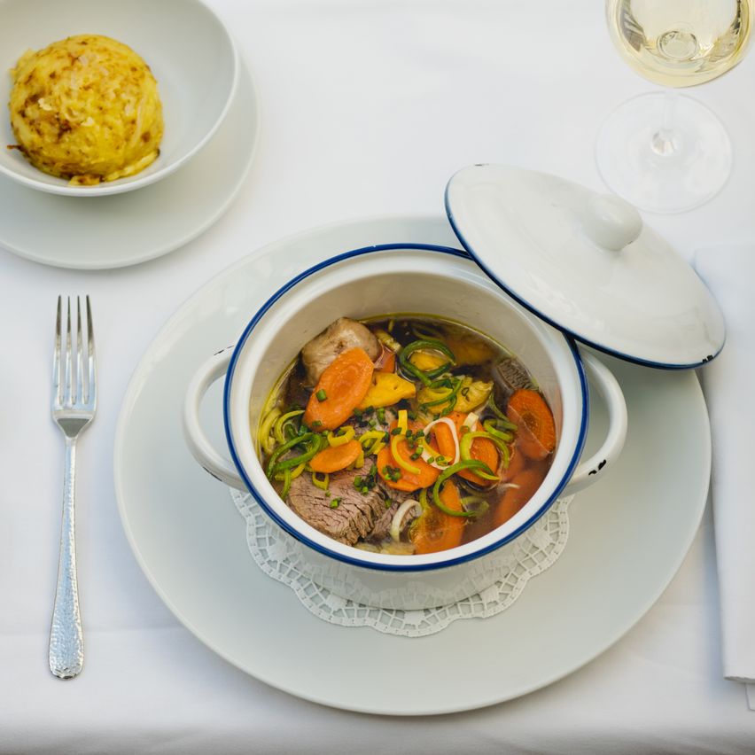 Ein Tisch mit einer Suppenschüssel, einem kleinen Teller mit einer gebratenen Kugel, einem Weinglas, einer Gabel und einem Serviettentuch auf einer weißen Tischdecke.