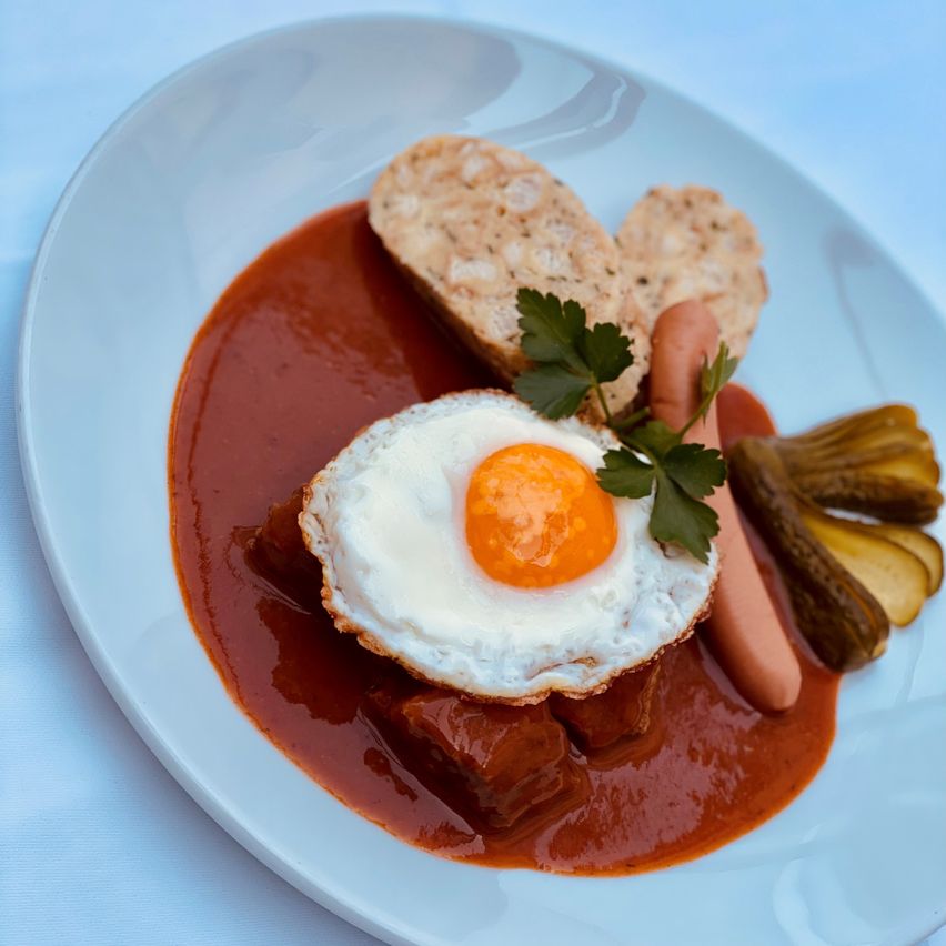 Ein Teller mit einem Spiegelei, roter Sauce, Brot, Gewürzgurken und einer Wurst auf einer weißen Tischdecke.