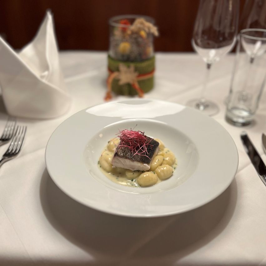 Ein weißer Teller mit einem Fischgericht und Garnierungen auf einem Tisch mit Weingläsern, Besteck und einer dekorativen Tischmitte.