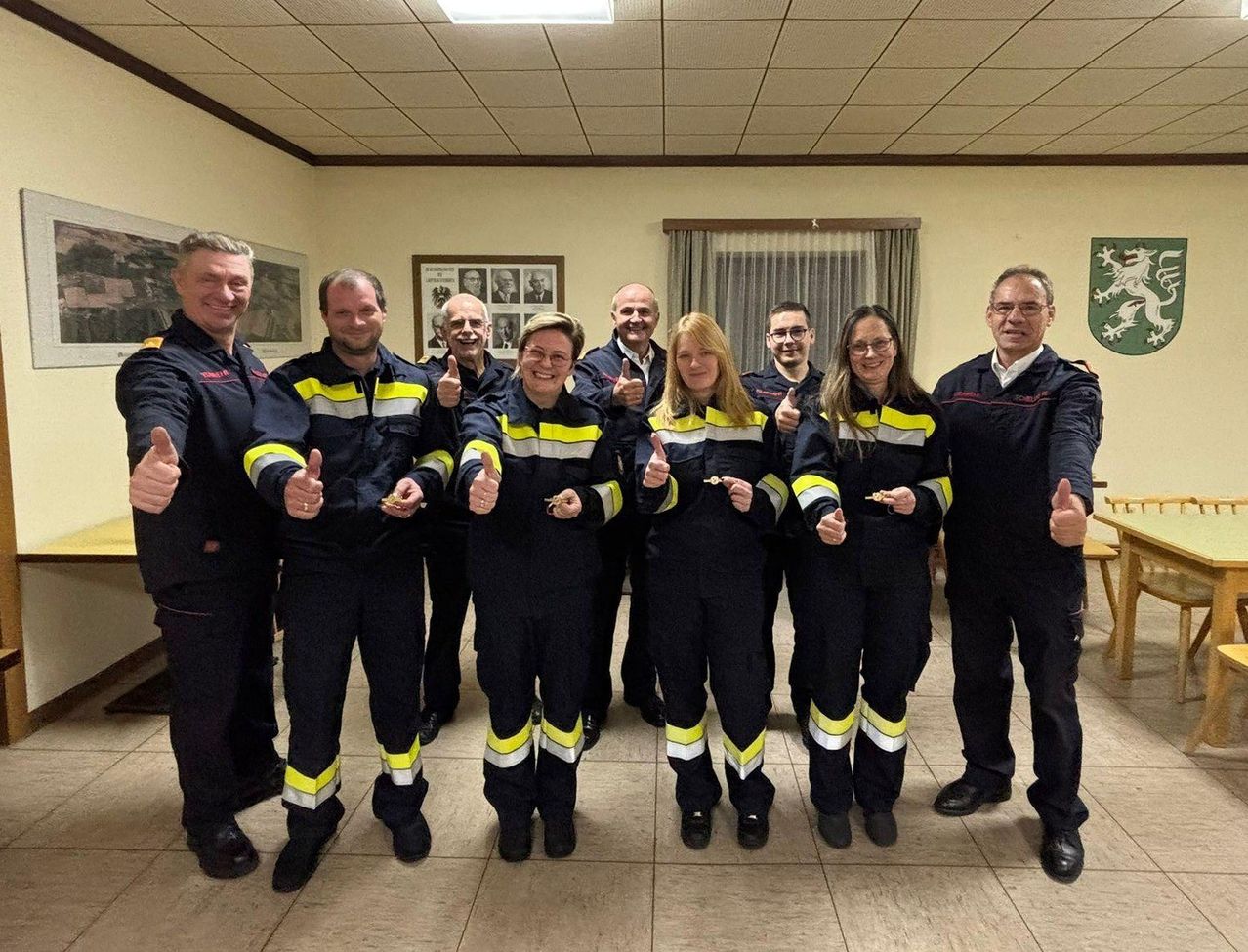 Eine Gruppe von Feuerwehrleuten in Uniform, lächelnd und Daumen hoch, stehen in einem Raum mit gefliestem Boden.