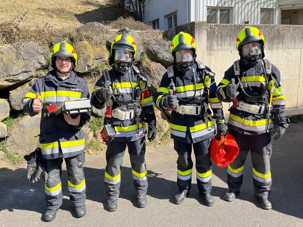 Vier Feuerwehrleute in voller Ausrüstung, einschließlich Helmen und Schutzanzügen, stehen zusammen draußen. Sie halten Ausrüstung in den Händen und scheinen einsatzbereit zu sein. Ein Gebäude mit Fenstern befindet sich hinter ihnen.