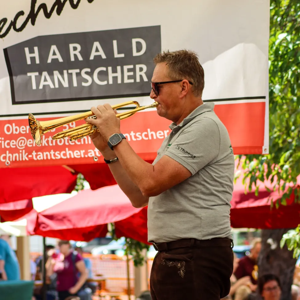Ein Mann spielt Trompete unter einem Banner mit dem Namen Harald Tantscher. Er trägt ein graues Shirt und braune Hosen.
