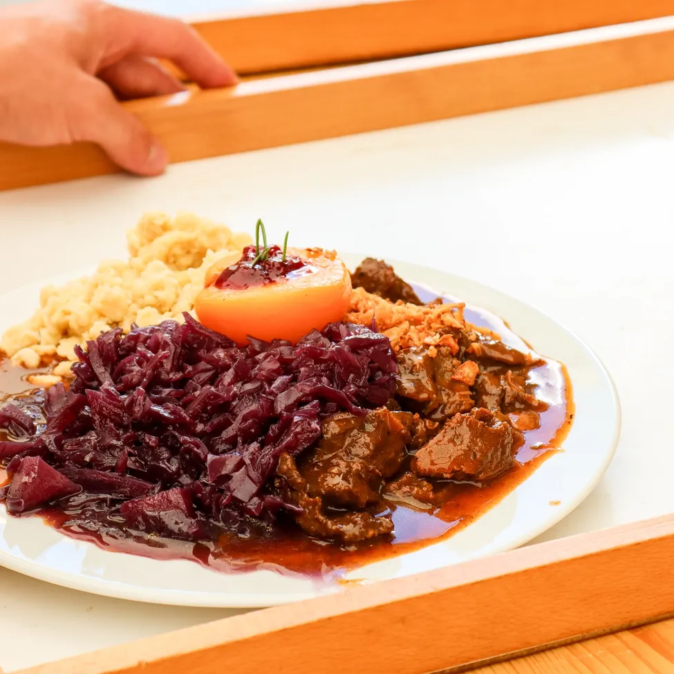 Ein Teller mit traditionellem deutschem Essen, bestehend aus geschmortem Rindfleisch, Rotkohl und einer Tomatenscheibe als Garnierung.
