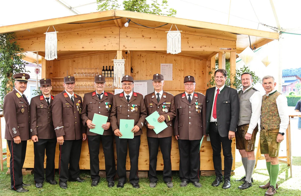 Sechs Männer in braunen Uniformen mit Medaillen stehen vor einem hölzernen Stand und halten grüne Papiere. Sie werden von einem lächelnden Mann in Anzug und zwei anderen in Freizeitkleidung umgeben.